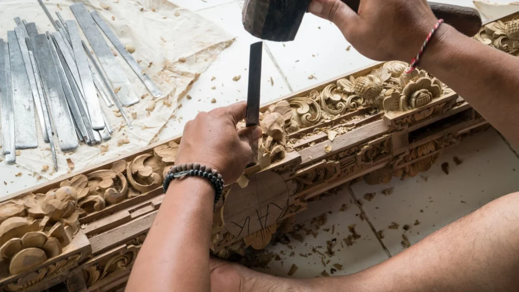 Personne sculptant des motifs floraux complexes dans du bois avec un ciseau et un maillet, des outils en métal à côté d'eux.