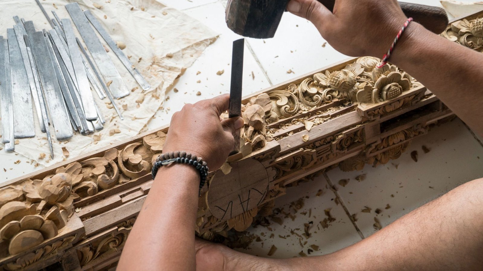 Personne sculptant des motifs floraux complexes dans du bois avec un ciseau et un maillet, des outils en métal à côté d'eux.