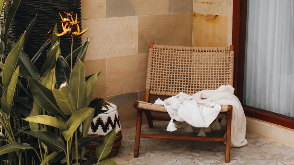 Une chaise tissée avec une couverture blanche est posée sur un patio en pierre, à côté de plantes en pot et d'un tabouret à motifs.