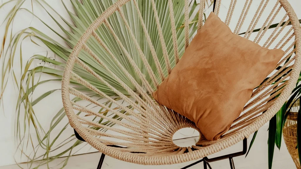 Une chaise en osier avec un coussin beige se trouve devant une grande feuille de palmier verte.