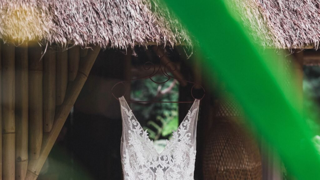 Une robe de mariée en dentelle est suspendue devant une structure en bois rustique au toit de chaume.