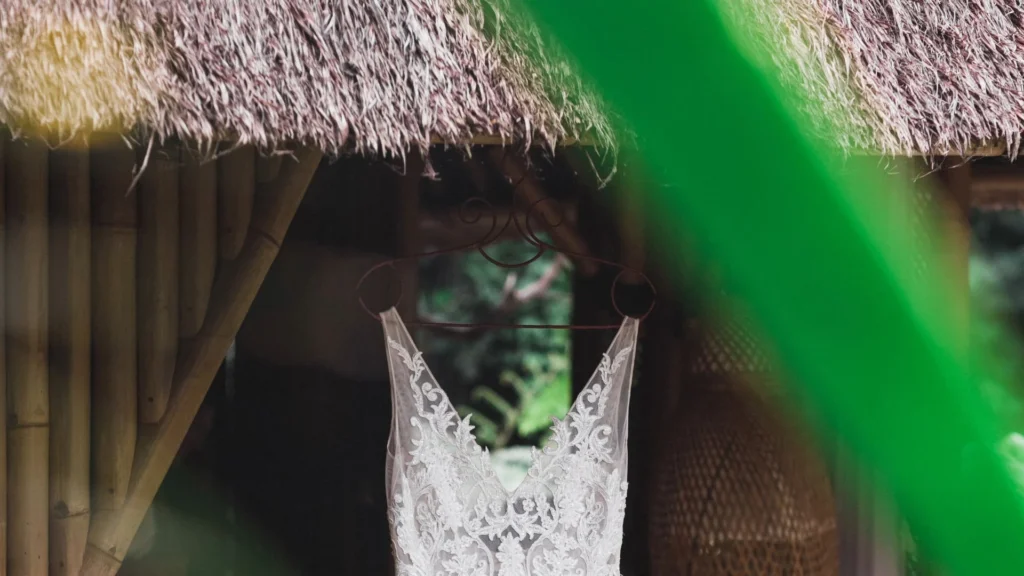 Une robe de mariée en dentelle est suspendue devant une structure en bois rustique au toit de chaume.