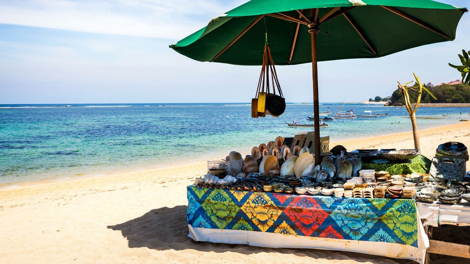 Stand de plage avec tissu à motifs colorés, exposant des coquillages et des bibelots sous un parasol vert près de l'océan.
