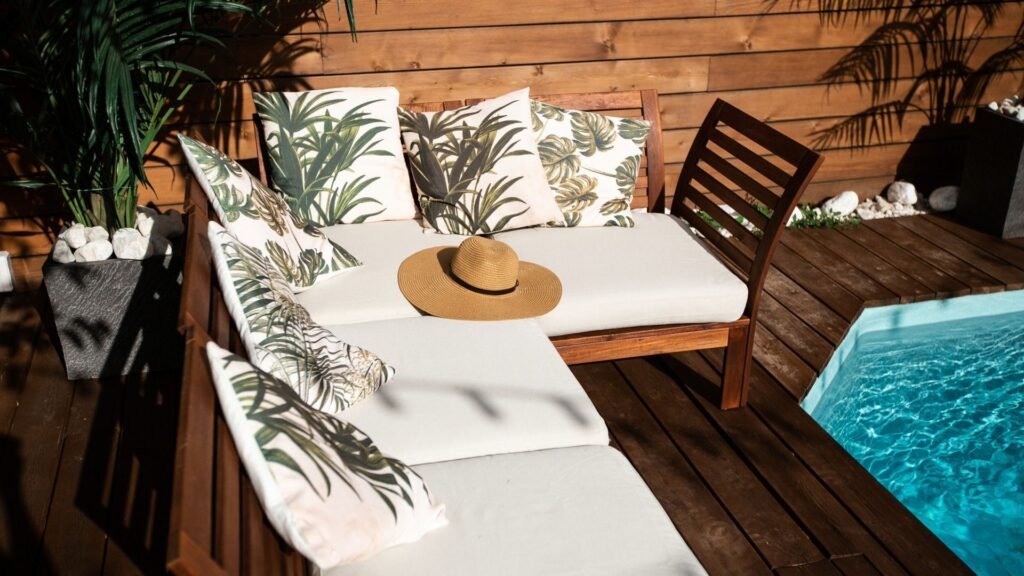 Terrasse extérieure avec un salon en bois, des coussins tropicaux, un chapeau à large bord et une petite piscine à proximité.