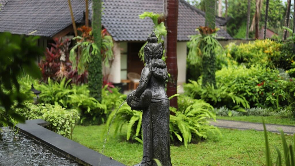 Statue d'une femme versant de l'eau dans un jardin luxuriant avec des plantes tropicales et une maison en arrière-plan.