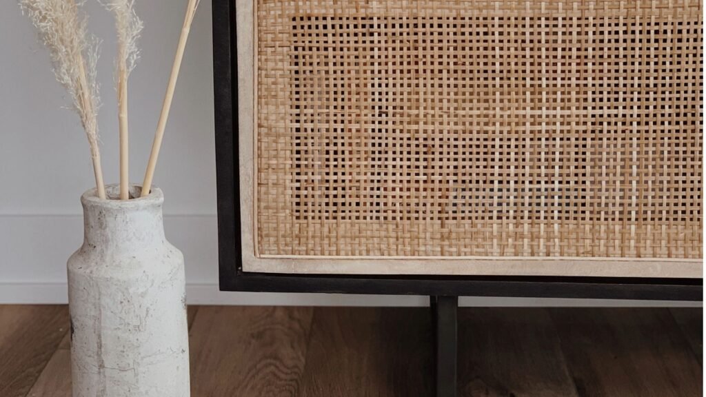 Décor minimaliste avec un vase blanc d'herbe de pampa et une armoire en rotin tressé sur un plancher en bois.