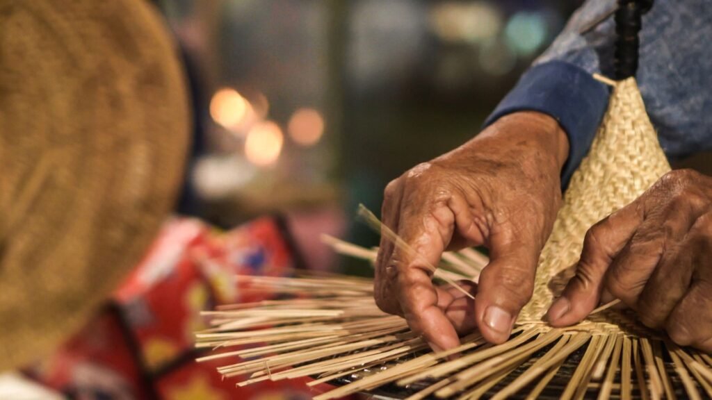 Des mains tissent de la paille pour en faire un chapeau, avec un arrière-plan flou et un éclairage chaleureux.