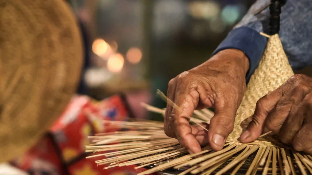 Des mains tissent de la paille pour en faire un chapeau, avec un arrière-plan flou et un éclairage chaleureux.