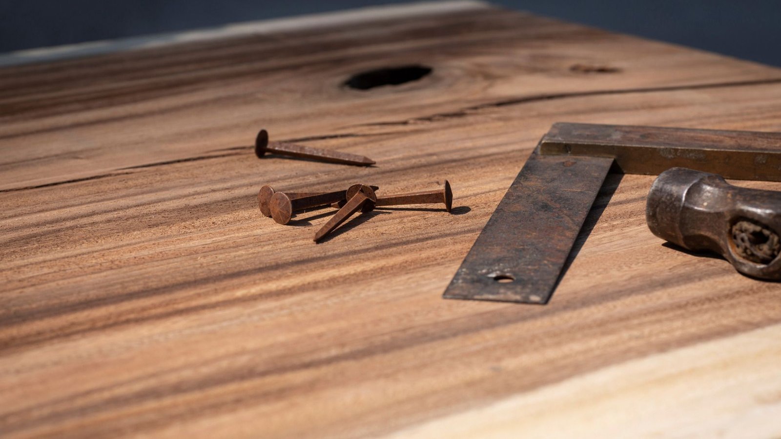 Des clous rouillés et un carré de métal placés sur une surface en bois avec un marteau à proximité.