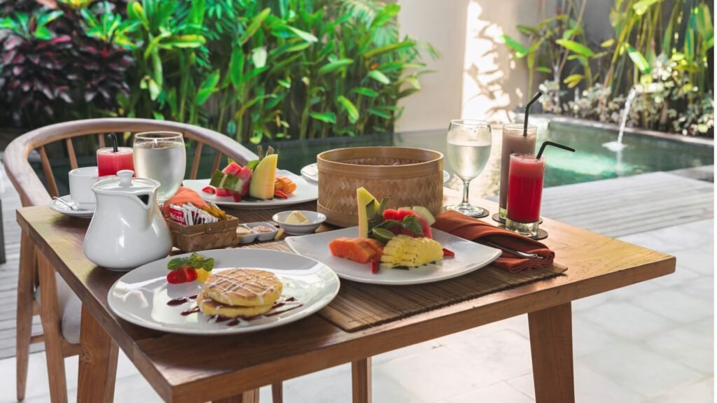 Petit-déjeuner en plein air composé de gaufres, de fruits et de boissons sur une table en bois près d'une piscine et d'une végétation luxuriante.